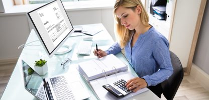 Young Businesswoman Calculating Bill In Office Young Businesswoman Calculating Bill With Computer And Laptop On Desk